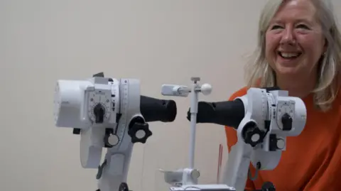 University Hospitals Sussex NHS Foundation Trust Joyce White, who was the head of service for orthoptics at the Brighton and Sussex University Hospitals NHS Trust. Joyce has blondish hair just below her shoulders and is wearing an orange shirt. She is sat beside orthoptic equipment.
