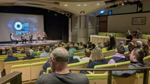 Oxford Shorts A group of people sitting in an auditorium during the Oxford Shorts Festival. Five people are on stage in front of a screen with the festival's name on.