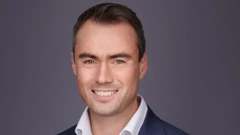YASA Tim Woolmer has short brown hair and is wearing a white shirt and blue blazer.