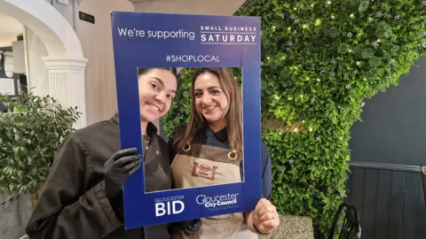 Gloucester BID Two women, with kitchen overalls on, are looking through an empty cardboard picture frame which says 'We're supporting Small Business Saturday' on the frame. They are standing in a restaurant with a large indoor green plant in the background
