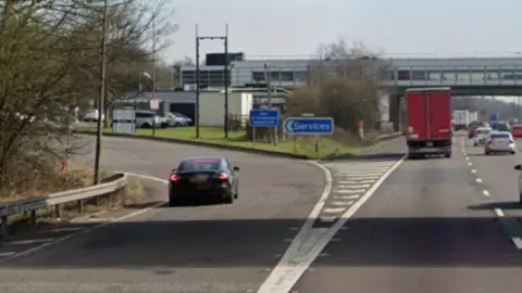 The slip road off to a motorway services, with lanes of traffic to the right, and a car driving around the bend to the services. There is a covered bridge over the lanes