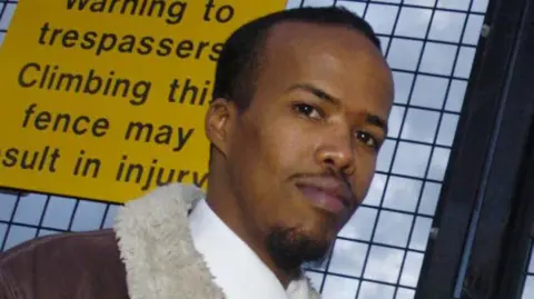 SWNS Essa Suleiman looks towards a camera with a yellow sign against his back 