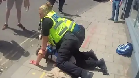 A still from a video of two police officers, both in uniform with high-vis vests are holding down a man on the pavement who has his arm out. People's legs are visible, indicating the public was aware and looking on.