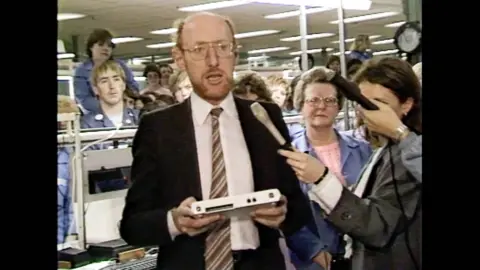 A man with a suit in glasses stands on the floor of a factory, surrounded by workers, holding a white home computer, while being asked questions by the press