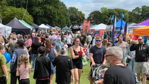 John Fairhall/BBC Lots of people are seen milling around a park in the sunshine. There are stalls in the background.