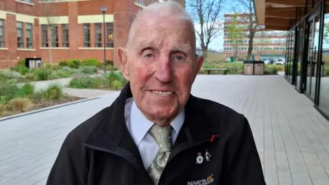 BBC/Seb Cheer An older man, Ken, looks into the camera and smiles. He is wearing a shirt and tie underneath a fleece and sits outside with a large brick building in the background.