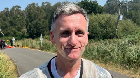 John Devine/BBC Wayne Swales has short grey hair. He is wearing a white shirt and a silver Hi-Vis jacket. He is standing on a road next to woodland.