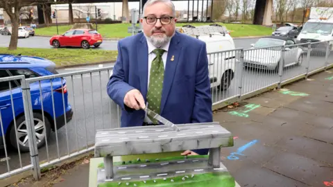LDRS Councillor Ron Beadle who has grey hair and is wearing a navy suit with a green tie. He is cutting a cake which is shaped like a road flyover. He is stood in front of Gateshead flyover. There are cars passing by behind him. 