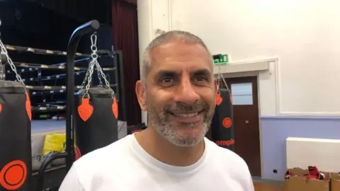 John Devine/BBC Paul Gill is wearing a white t-shirt. He has cropped grey hair and a beard and moustache. Behind him is a boxing ring on a stage. Punch bags hang on stands on the floor below.