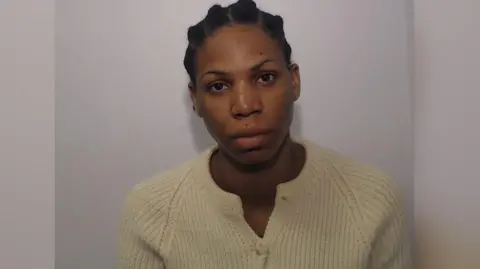 Greater Manchester Police Ruth Auta looks straight at the camera in this police photo. She has braided hair which is tied back and is wearing a cream coloured cardigan. There is a Greater Manchester Police logo in the top left hand corner.