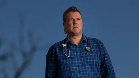 The Age/Jason South Dr Chris Webster standing outside, a bare tree in the foreground. A stethoscope is around his neck and he is wearing a blue check shirt.