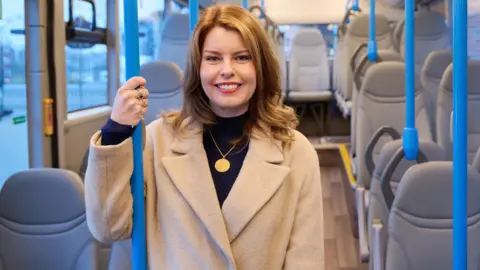 North East Mayor Kim McGuinness. She has shoulder length brown hair, a beige coat, black top and is wearing a gold necklace and two rings. She is holding on to a metal pole on the bus and is smiling. There are empty, grey seats on the bus.