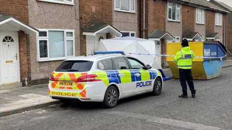 A police officer and a police car was parked next to a terraced house, which has been closed. A white tent is in front of the door area. There is also a yellow and blue metal skip on the right side of the image.