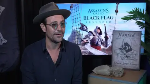 Ubisoft Matt Ryan is sitting on a chair wearing a hat and glasses, speaking to someone on the other side of the camera. Behind him is promotional material for the upcoming Assassin's Creed Black Flag game.