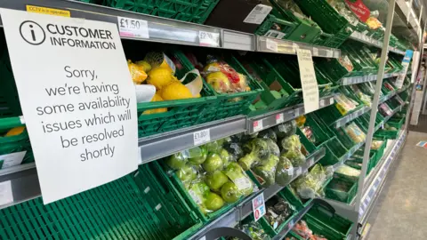 Empty green food crates that should be full of fresh fruits and vegetables on sale at the Co-op Food shop in Brundall. 
Customer information sign in the foreground which reads "Sorry we're having some availability issues which should be resolved shortly" 