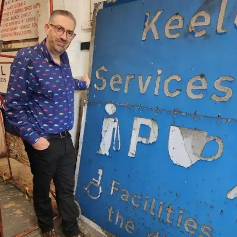 National Highways A man in a blue shirt with colourful prints stands with his hand on the blue Keele Services sign, which is propped against a wall