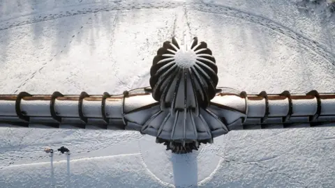 PA Media People walking in the snow at the foot of Antony Gormley's Angel of the North sculpture in Gateshead. The tall sculpture, which resembles a man with rectangular wings, is dusted with snow.