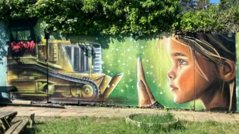 Shariqua Ahmed/BBC Mural of a girl holding up a hand to a yellow bulldozer- with a green background painted on a wall lined with trees
