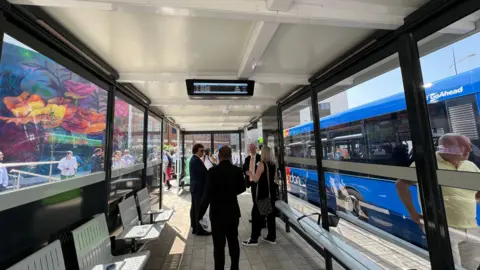 A group of people dressed smartly stand inside a new bus shelter, which is covered on all sides. Through the glass, a Swindon bus can seen, which is painted blue