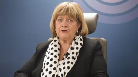 PA Media A woman with shoulder length blonde hair and a fringe. She is wearing a black blazer and a white and black polka dot scarf.