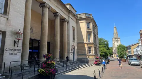 The front entrance to the Gloucestershire Shire Hall has four pillars in front of it and some flowers on the side. There are people walking past it on the street in front.