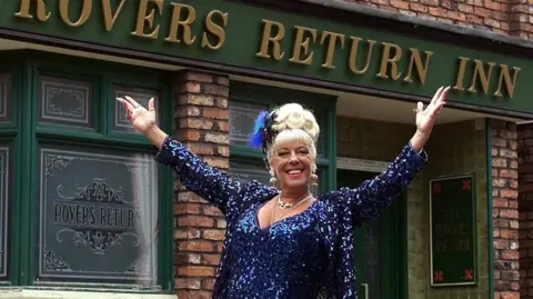 PA Julie Goodyear, blonde hair up, dressed in sparkles and heavy make-up outside Coronation Street's Rovers Return Inn.