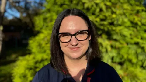 Elliot Deady/BBC A smiling woman looking at the camera, standing in front of a bush in a garden.