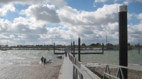 Brightlingsea town jetty with beach in the foreground, two people launching a boat on the left, others standing on the jetty with a boat, water, moored boats and trees in the distance under white fluffy clouds with blue sky glimpsing through