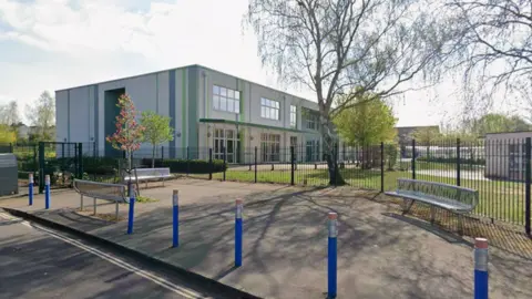 A screenshot from Google street view showing the exterior of May Park Primary School. It is a grey-white rectangular building with green and blue stripes and large windows. There is a small green space with trees growing in front of the building. Outside the metal fencing, there are blue bollards shaped to look like pencils spaced out evenly on the pavement.