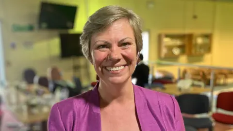 Woman wearing pink blouse smiling for the camera with a blurred background of tables and chairs
