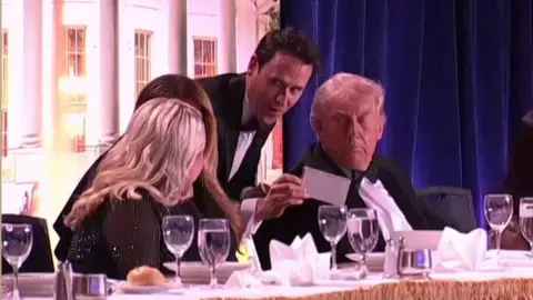 Speaker Oz Pearlman (standing in the centre with a piece of paper) talks to US President Donald Trump (right), First Lady Melania Trump (2nd left) and White House press secretary Karoline Leavitt (left) at the White House Correspondents Dinner in Washington DC. Photo: 25 April 2026