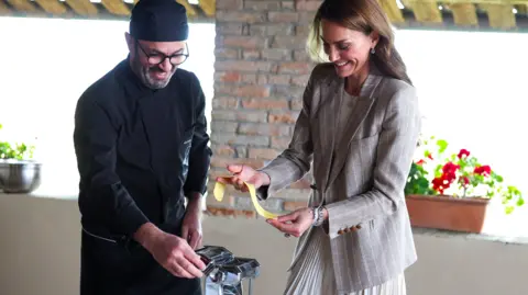 PA Media An Italian chef dressed in all black kitchen wear and hat stands to the left of the image next to the Princess of Wales as he helps her make fresh pasta in a kitchen. The Royal, dressed in a grey blazer and skirt is grinning as she holds the pasta in her hands