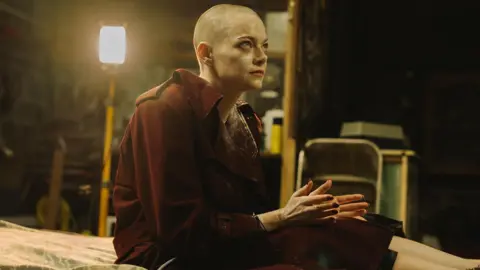 Universal Emma Stone in Bugonia, with handcuffs on and a shaved head