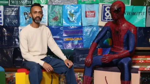 Mr Patel is sat down next to a life-size Spiderman model wearing a red and blue costume with a black spider on the front. Mr Patel is wearing jeans and a cream jumper. Behind them are rows of plastic bags with company logos on