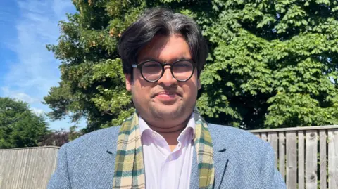 A man with black hair and black round spectacles standing outside in front of a tree on a bright, sunny day. He is wearing a light blue blazer and a green tartan scarf.