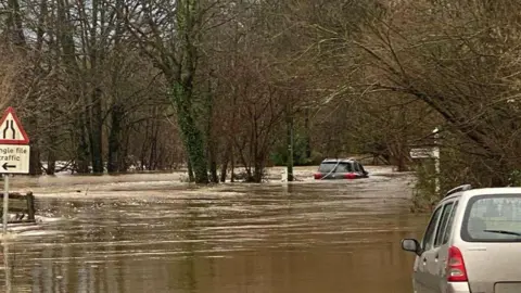 A dark grey car is partially submerged in brown flood water. There is a light grey car in the foreground to the right of the image.  There are trees and street signage around.