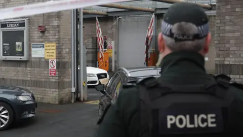 A police officer stands with his back to the camera and is stood near a grey, unmarked police car. There is a police cordon closing off the station and a white Audi car is visible in the distance in the station, flanked by two large grey brick blast walls.