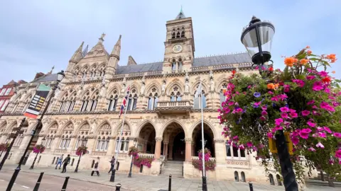 Martin Giles/BBC Outside of the Guildhall in Northampton. On the right of the picture is a hanging basket of pink and orange flowers. The sky is cloudy.