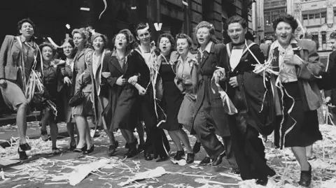 Men and women are lined up singing and dancing and covered in streamers as celebratations are held on VE day in this black and white archive photograph. The women are wearing 1940s skirts, jackets and blouses and two of the men are in uniform. There are reams of streamers underfoot.