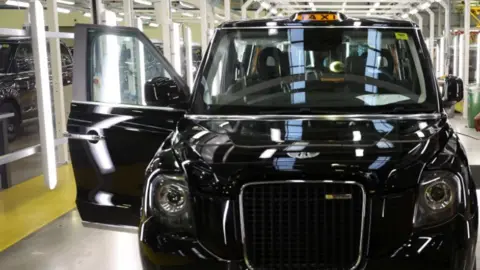 A modern black cab is pictured on a production line. It is shiny, and one of the doors is open