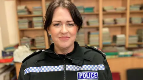 Zoe Kelsey is wearing a police uniform and the word police is on the right hand side of her jacket. She has brown hair tied back. 