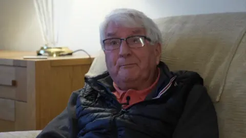 A man who appears to be in his sixties or early seventies, Stuart Couzens. He has wispy white hair which falls across his forehead. He wears thin framed, black glasses. He has a kind looking face. He wears a black gilet, dark grey jumper, and coral polo neck shirt.