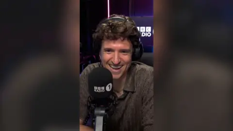 Greg James has wavy brown hair and is wearing a dark olive green shirt with a V-neck and collar.