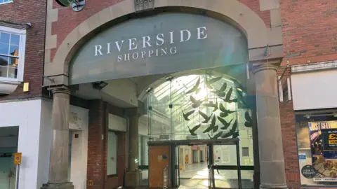 The entrance to a shopping centre which features large glass doors and a stone archway. A sign above the door has white lettering indicating it it the Riverside shopping centre.