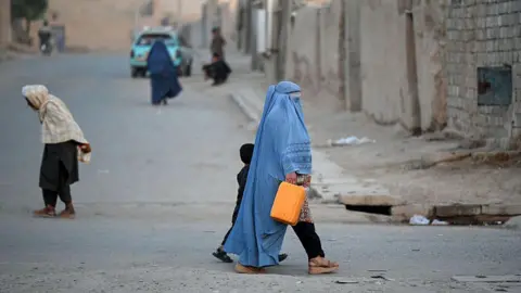 An Afghan burqa-clad woman walks along a street in the 7th district of Afghanistan's Kandahar, several people are behind her. She is walking next to a child. The street is dusty and beige.
