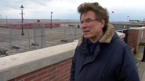 Daren Hale has short brown windswept hair and is wearing rimless spectacles on his face and a navy blue coat. He is standing next to a redbrick wall, which is in front of tall safety barriers around the perimeter of the pier. 
