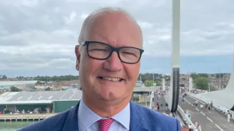 Laura Foster/BBC Matthew Hicks is standing high above the Gull Wing bridge in Lowestoft. He is wearing a blue suit, blue shirt and pink tie. He is wearing glasses and smiling. 