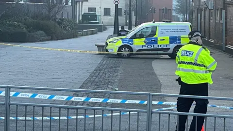 A photo of a police cordon in Willow Row, Derby. Police tape can be seen across a walkway, and a police officer with his back turned is stood next to the tape. In the distance, on the other side of a the cordon, a police van is parked. 