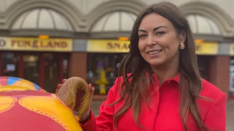 Serena Silcock has long brown hair, she is wearing a red jacket and gold earing and is stood outside Southport Funland