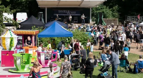 A small summer festival on a sunny day. Crowds of people including parents with prams sit or stand on a patch of grass in front of a stage where a band is performing. A spinning teacup ride is visible in the foreground. 